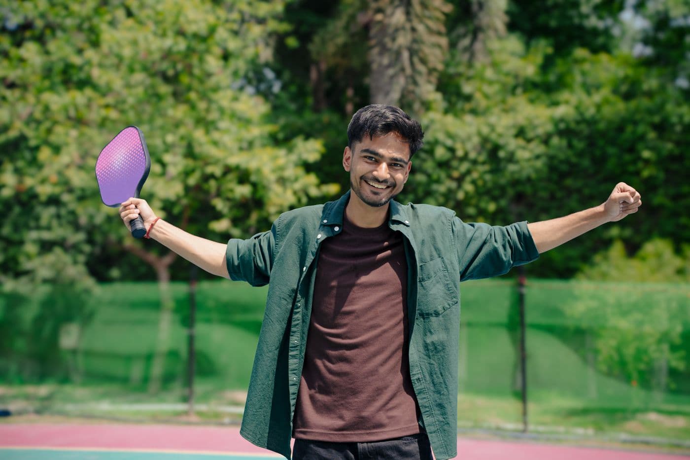 A player on an outdoor court holding a pickleball paddle up, arms wide, mid-celebration.